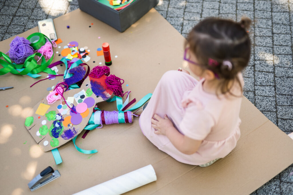 Dziewczynka siedzi na kartonie leżącym na ziemi. Na kartonie leży lalka zrobiona z kartonu. Materiały i przybory.