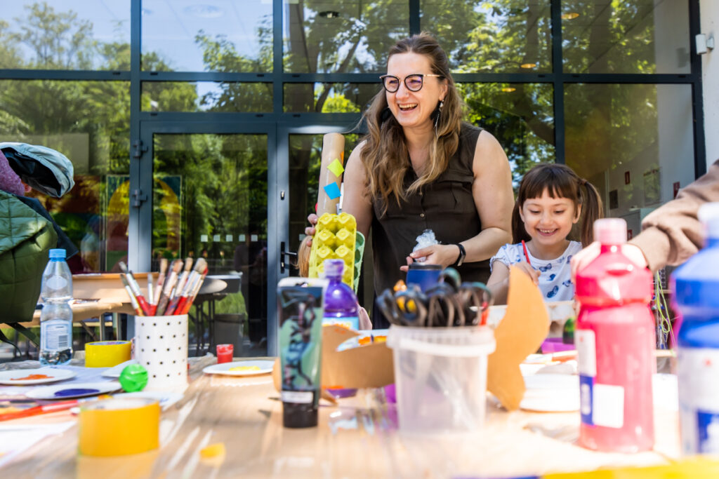 Uśmiechnięta kobieta w okularach. Obok niej uśmiechnięta dziewczynka. Przed ni stół, na którym stoją materiały do prac plastycznych.