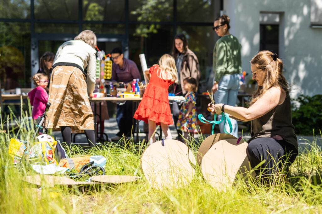 Na przedzie kobieta w okularach kuca. W rękach ma nożyczki i kolorową taśmę. W tle grupa dzieci i dorosłych przy stole.