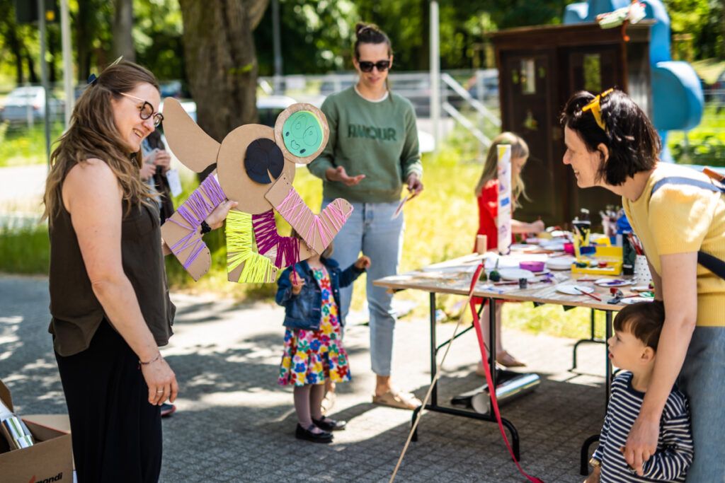Kobieta w okularach trzyma w rękach fantastyczną kartonową postać. Przed nią stoi kobieta z dzieckiem i przyglądają się.