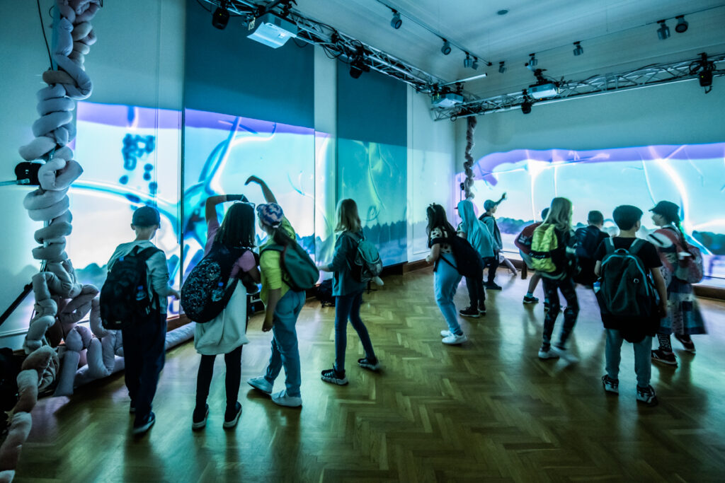 W sali na dwóch ścianach wyświetlane są fantastyczne, abstrakcyjne obrazy. Grupa dzieci przygląda się im.