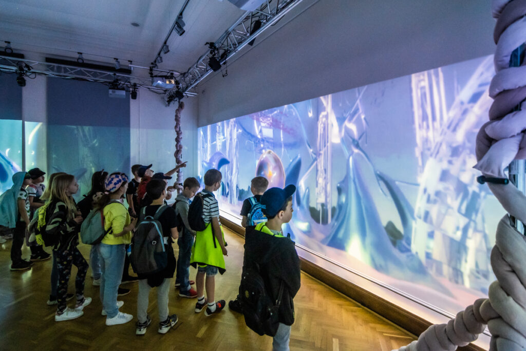 W sali na dwóch ścianach wyświetlane są fantastyczne, abstrakcyjne obrazy. Grupa dzieci przygląda się im.