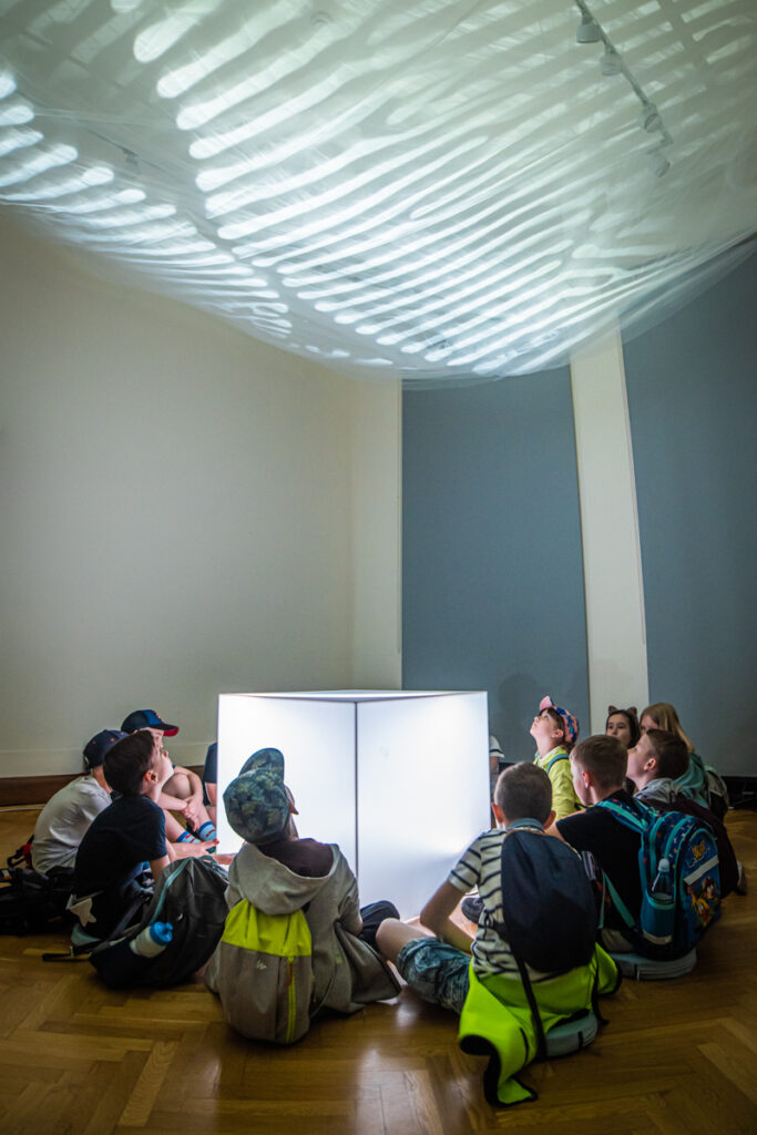 Na środku sali lezy duży, podświetlony sześcian. Wokół niego siedzi grupa dzieci. Spoglądają na sufit, na którym wyświetlane są abstrakcyjne wzory.