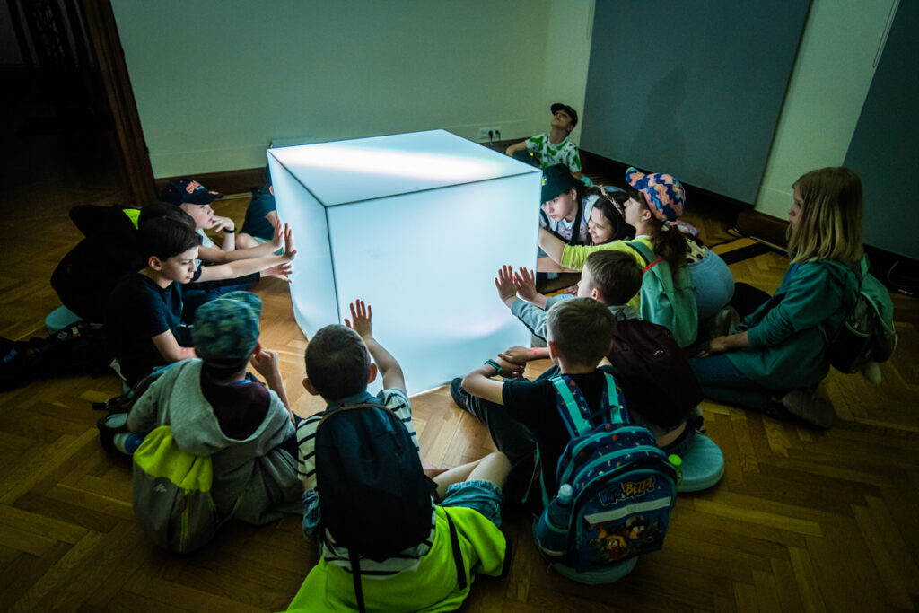 Na środku sali leży duży, podświetlany sześcian. Grupa dzieci siedzi na około. Dotyka sześcian rękami.