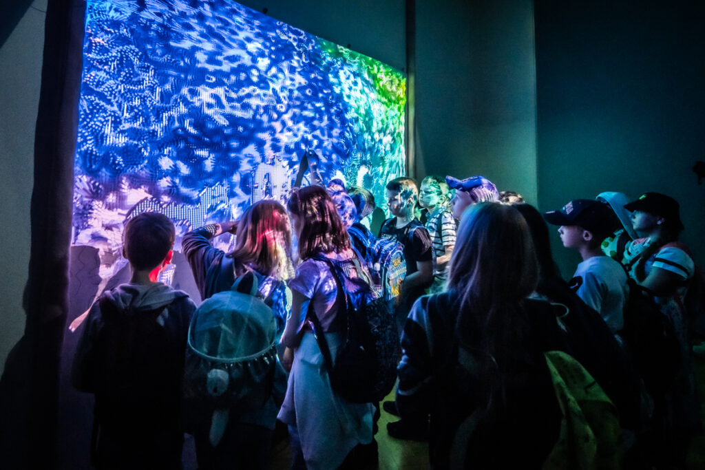 W sali po lewej stronie duży ekran, na którym wyświetlana kolorowa projekcja. Przed ekranem grupa dzieci. Część dotyka ekranu.