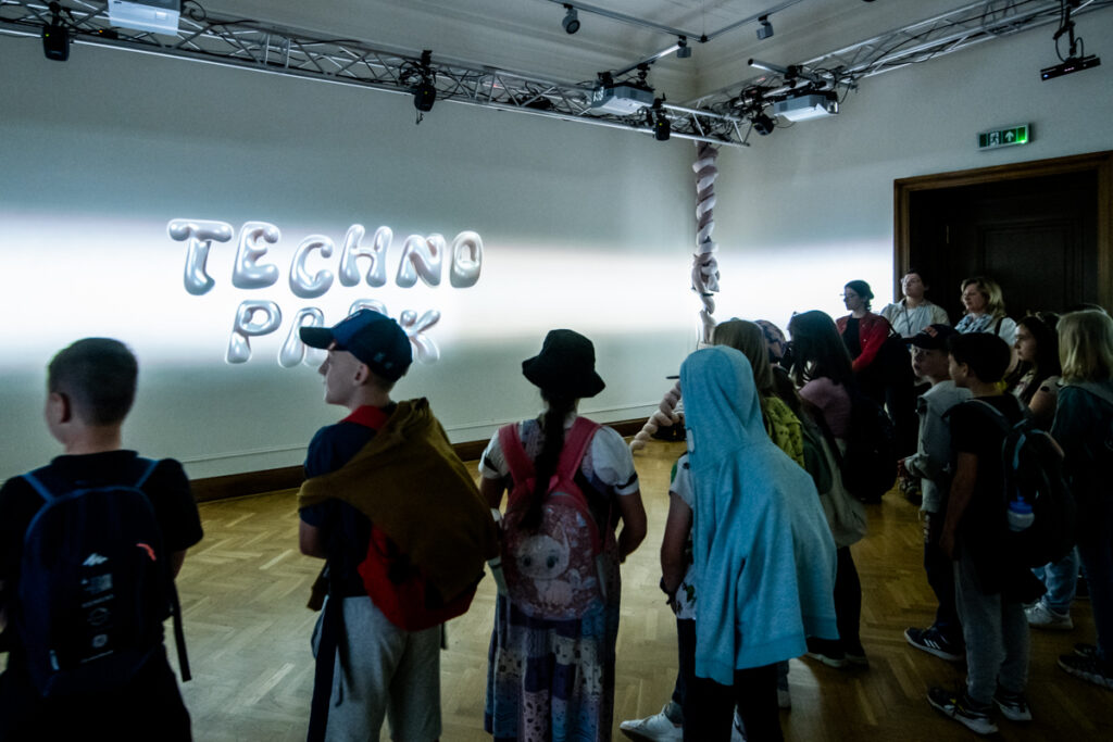 W sali na ścianie wyświetla się napis: Techno park. Liczna grupa dzieci i dorosłych stoi.