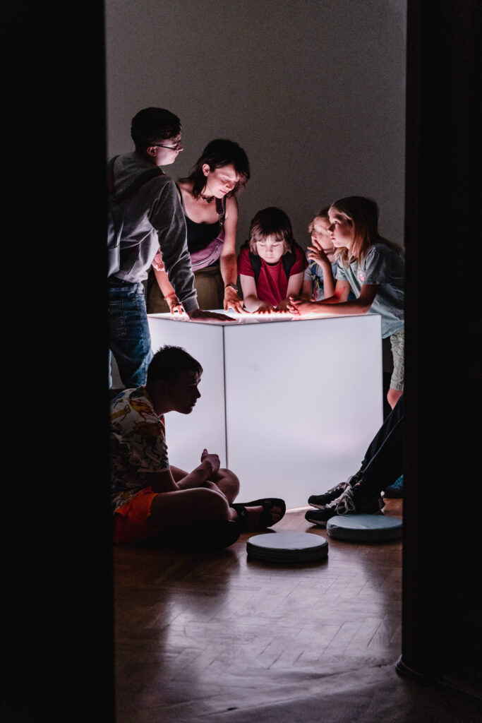 Na środku leży duży, podświetlany sześcian. Grupa osób stoi nad nim. Część się pochyla na nim, inni się opierają rękoma Chłopiec siedzi obok sześcianu z przodu.