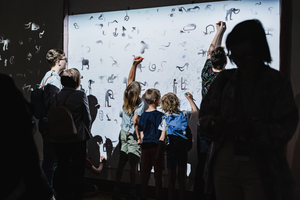 Na ścianie w tle wyświetlany jest ikonograficzny ornament. Kilkoro dzieci stoi przed nią i rysuje na niej.