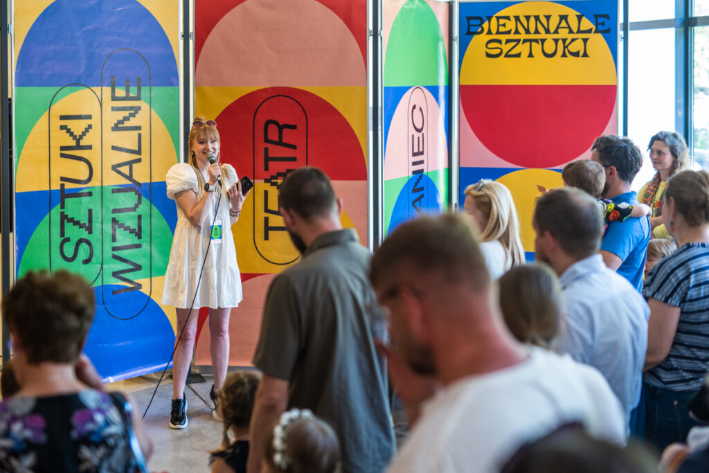 Pracownica CSD stoi przed grupą dzieci i dorosłych. Mówi do nich. Za nią kolorowe plakaty