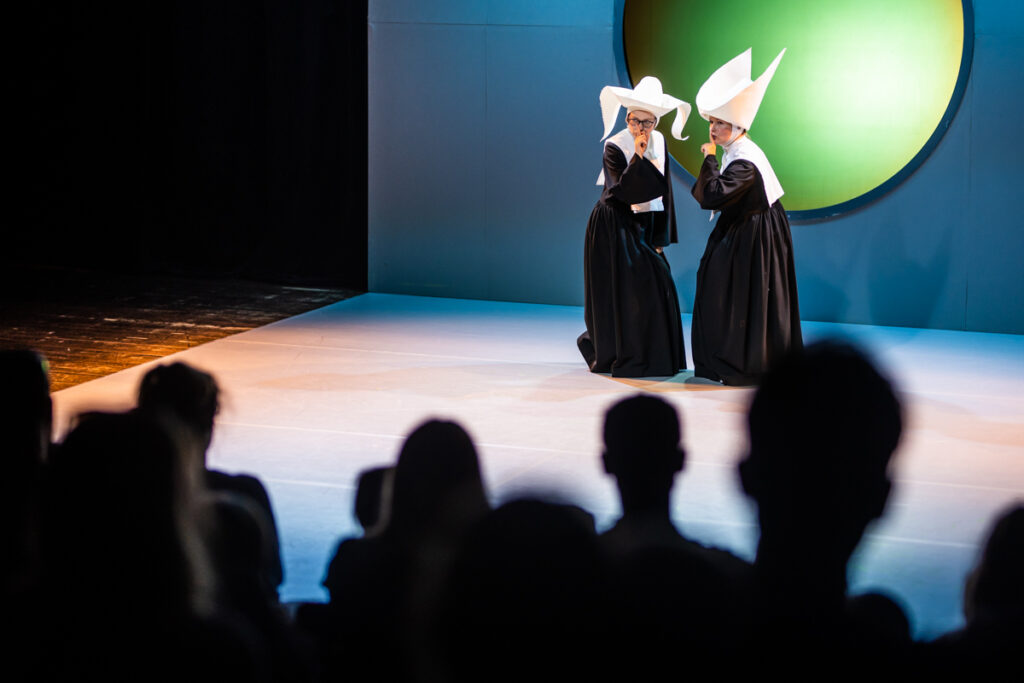 Na przedzie widownia. W głębi scena, na której stoją dwie aktorki ubrane w strój przypominający habity. Z wielkimi kapeluszami.