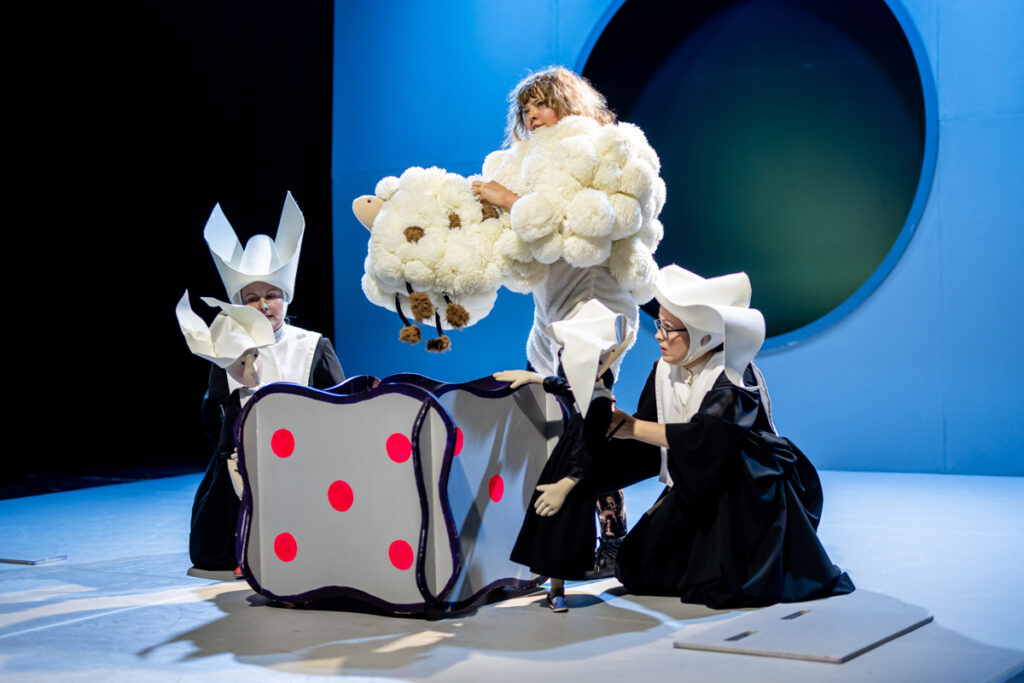 Na scenie. Na środku stoi duży otwarty pojemnik. Jego boki przypominają kostkę do gry. Za pojemnikiem stoi aktorka ubrana w puchowy stój, w rękach ma kukłę owcy, w tym samym kolorze co jej stój. Obok pojemnika siedzą dwie inne aktorki. Ubrane są w habit. W rękach mają kukły, które są ubrane w takie same habity.