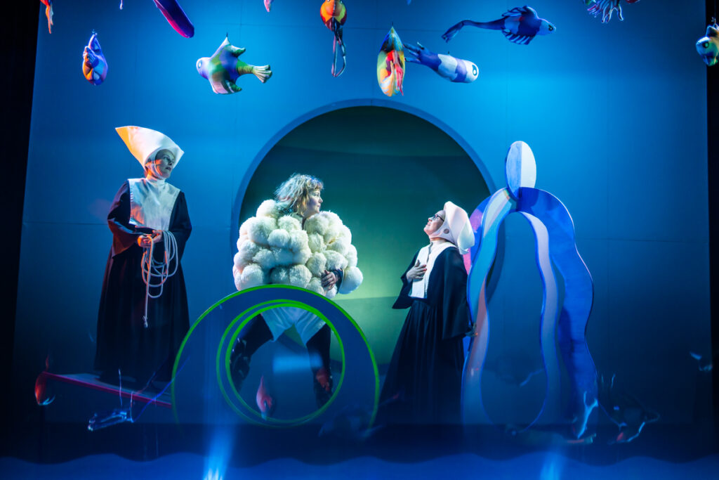 Na scenie Trzy aktorki. Aktorka stojąca pośrodku ubrana jest w puchaty kostium. Dwie po jej po bokach mają na sobie habity zakonne. Nad ich głowami na sznurkach wiszą kolorowe ryby.