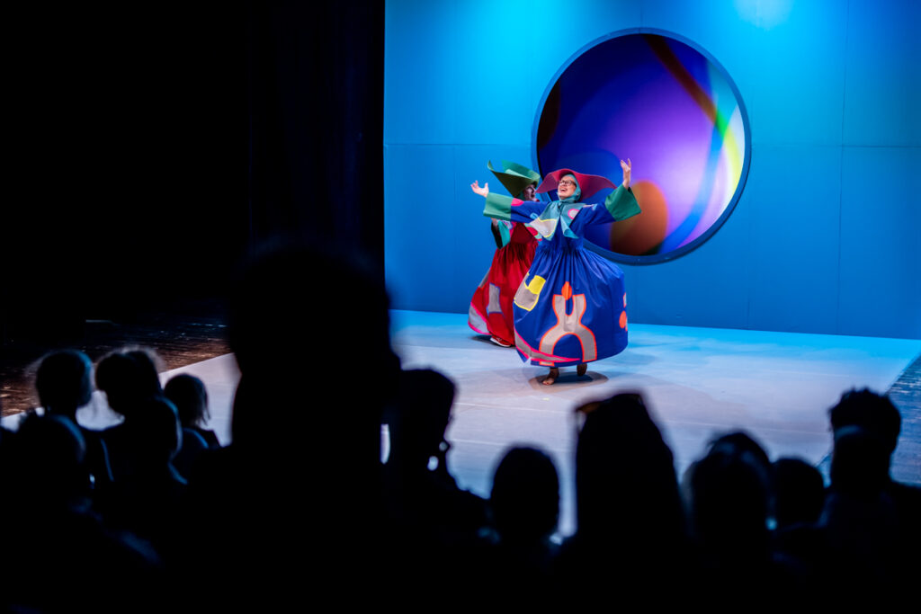 Widownia. W tle scena. Na scenie dwie aktorki ubrane w kolorowe, długie suknie i wielkie kapelusze. Tańczą.