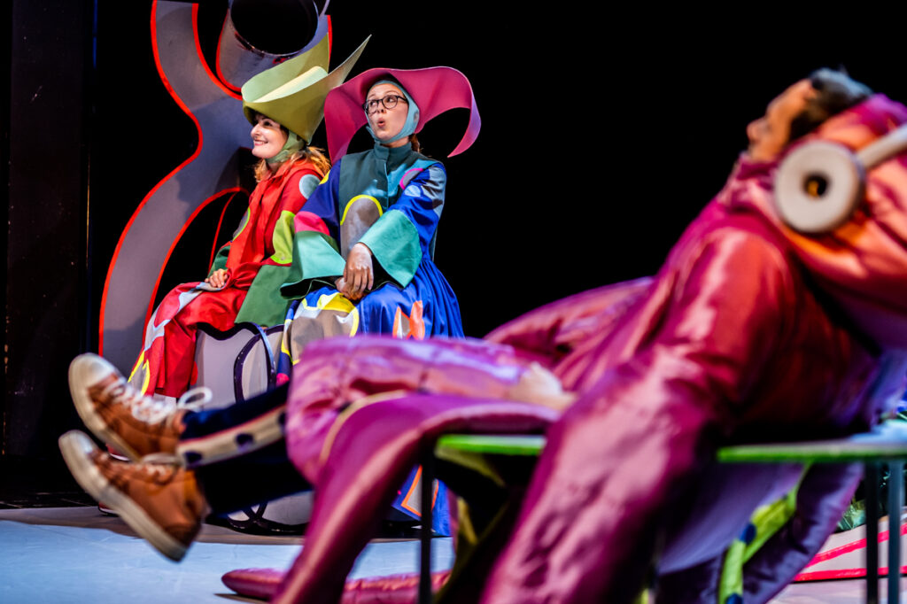 Scena teatralna. Z przodu aktor ubrany w połyskujący kostium ośmiornicy. W tle dwie aktorki ubrane w kolorowe suknie i wielkie kapelusze. Jedna z nich uśmiecha się. Druga gwiżdże.