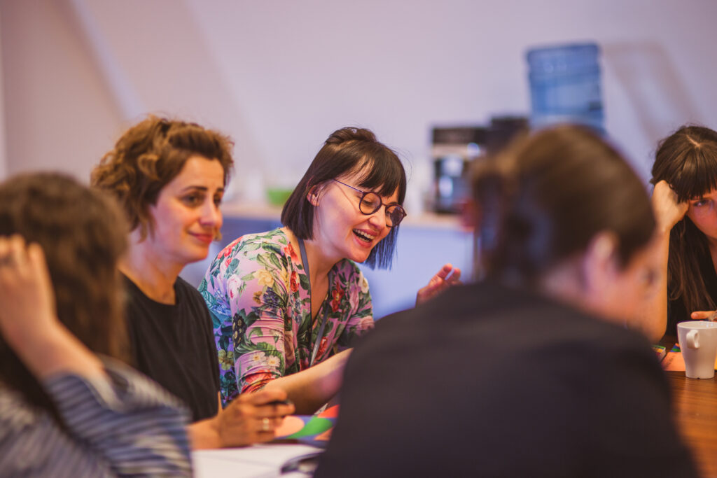 Warsztaty. Kilkoro osób siedzi przy stole. Kobieta uśmiecha się radośnie.