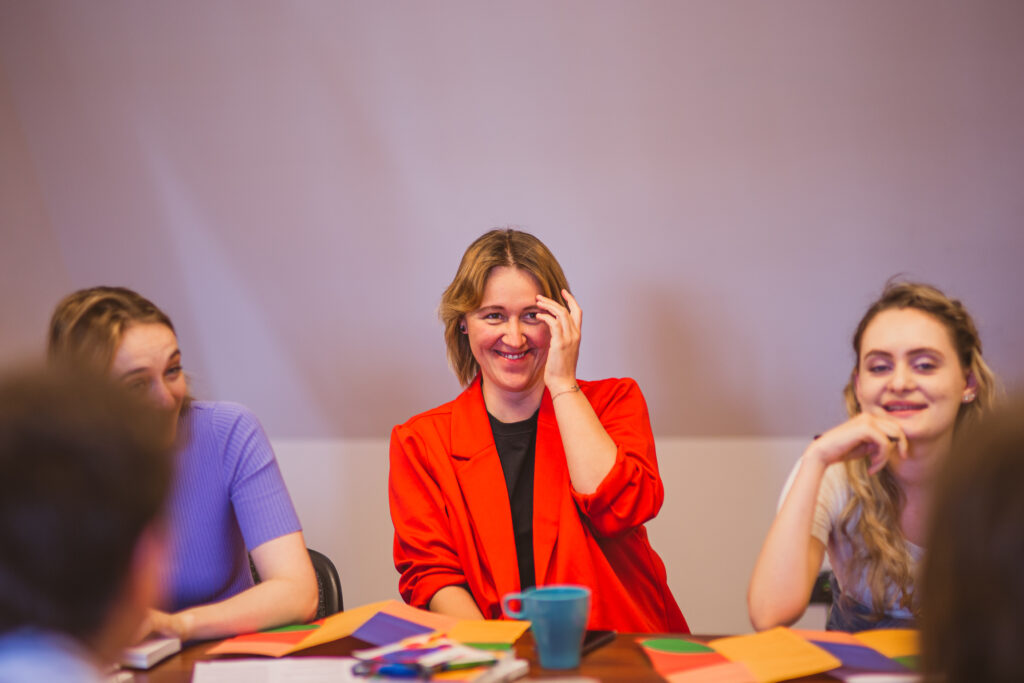 Warsztaty. Trzy kobiety siedzą przy stole i się uśmiechają. N stole leżą ulotki.