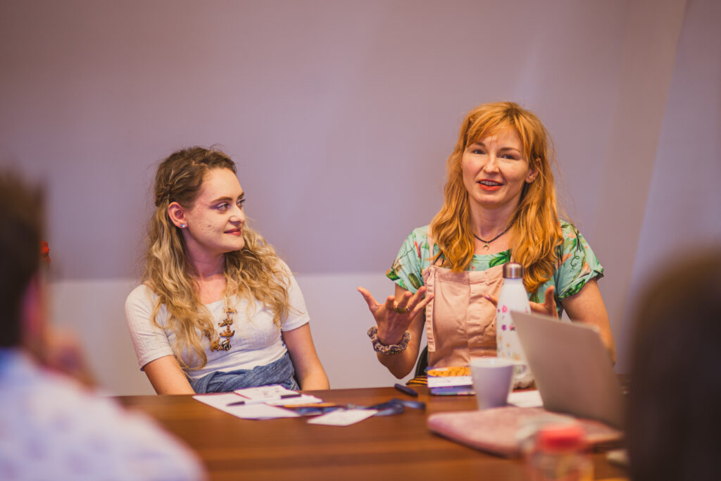 Warsztaty. Dwie kobiety siedzą przy stole. Jedna opowiada o czymś. Druga patrzy na nią uważnie.