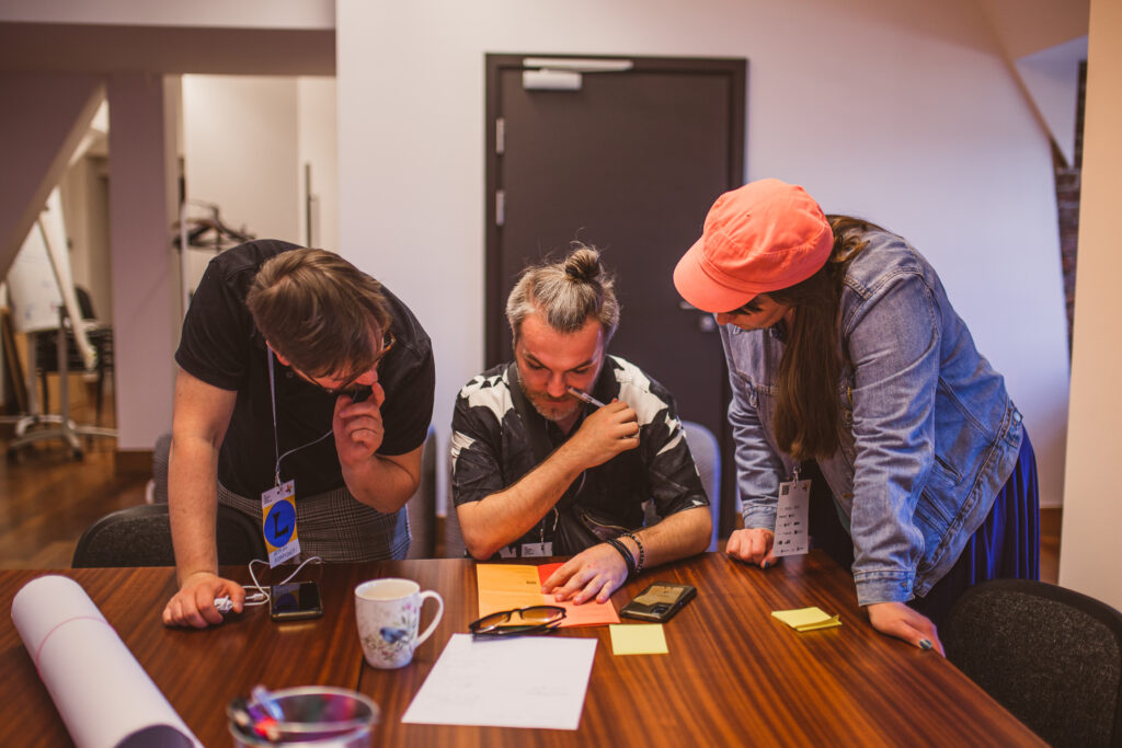 Warsztaty. Mężczyzna siedzi przy stole. Dwie inne osoby pochylają się nad nim. Wszystkie przyglądają się informacjom z ulotki.