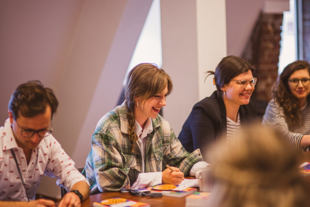 Warsztaty. Kilkoro osób siedzi przy stole. Uśmiechają się. Mężczyzna po lewej notuje.