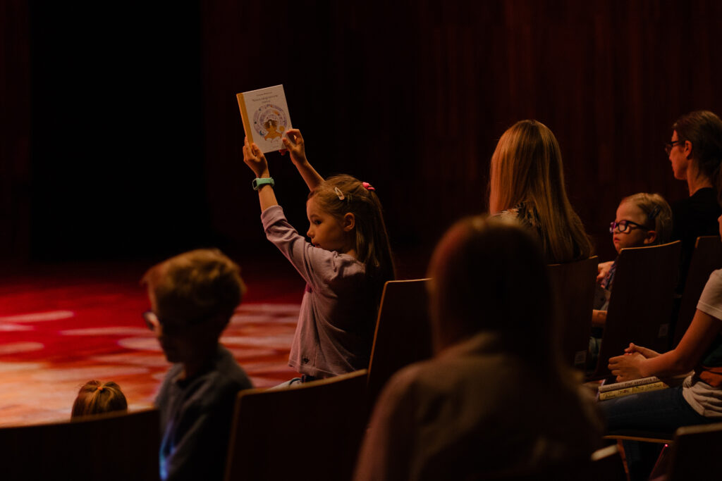 Widownia. Dziewczynka na przedzie trzyma w rękach książkę i unosi ją wysoko.