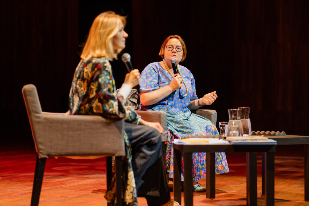 Dwie kobiety siedzą na scenie. Mają w rękach mikrofon.