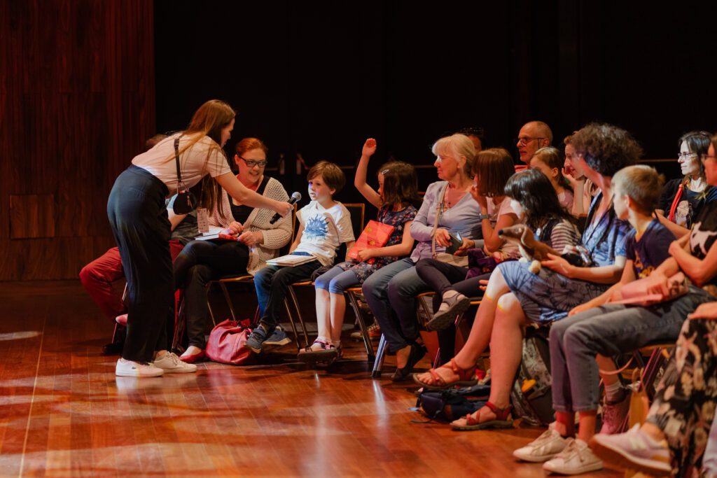 Widownia. Grupa dzieci i dorosłych. Kobieta z mikrofonem podeszła do chłopca. Chłopiec mówi coś.
