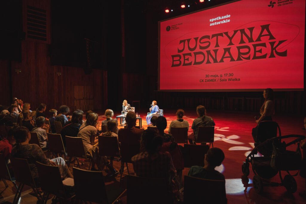 Na widowni grupa osób. W tle na scenie siedzą dwie kobiety. Za nimi duży ekran, na którym wyświetlany jest napis: Spotkanie autorskie Justyna Bednarek.