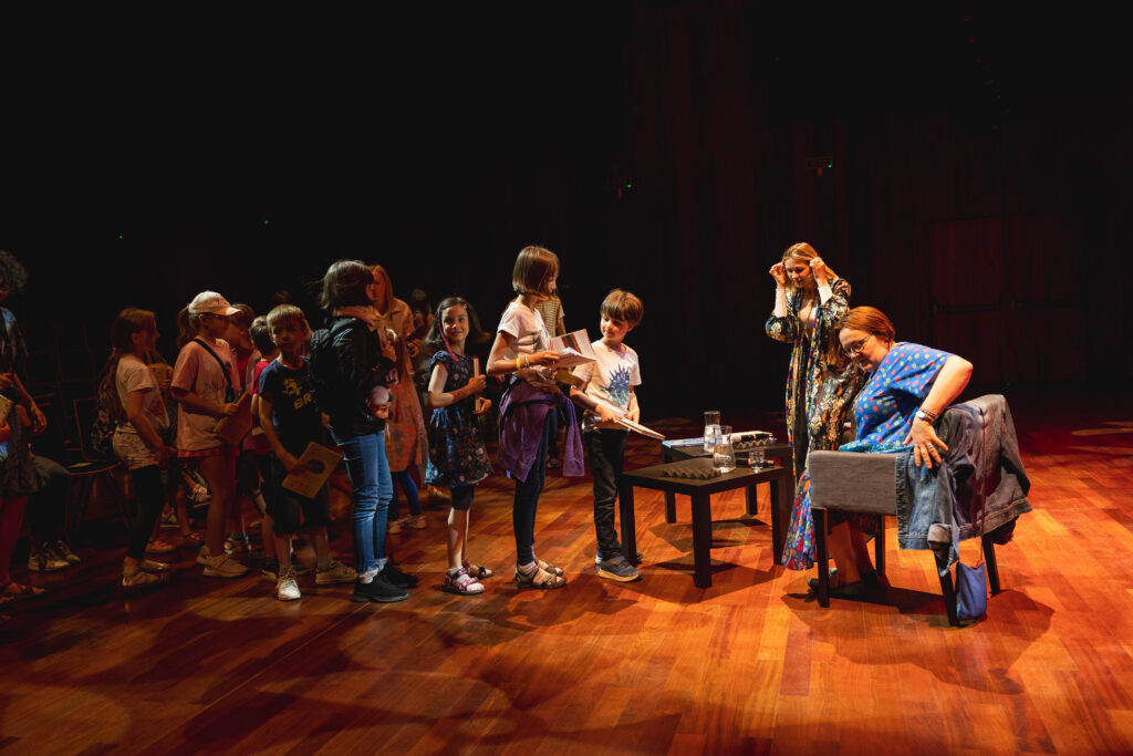 Po lewej widownia. Na scenie po prawej kobieta siedzi w fotelu. Przed się ustawione w kolejce stoi grupa dzieci z książkami w rękach.