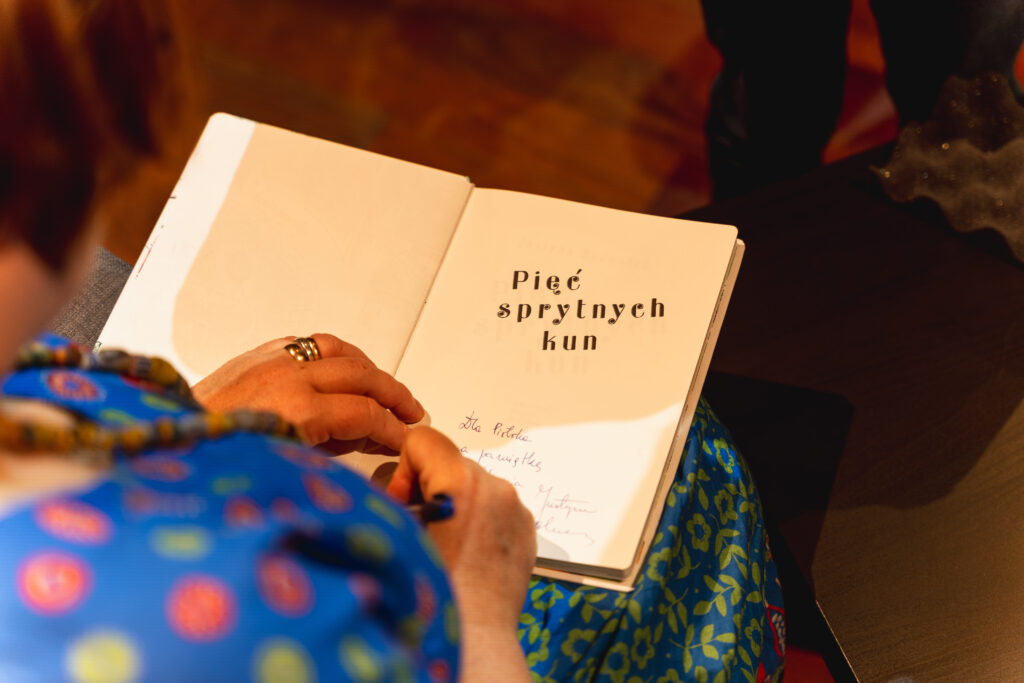 Kobieta trzyma na kolanach otwartą książkę. Pisze na stronie tytułowej adnotację. Widać tytuł: Pięć skrytnych kun.