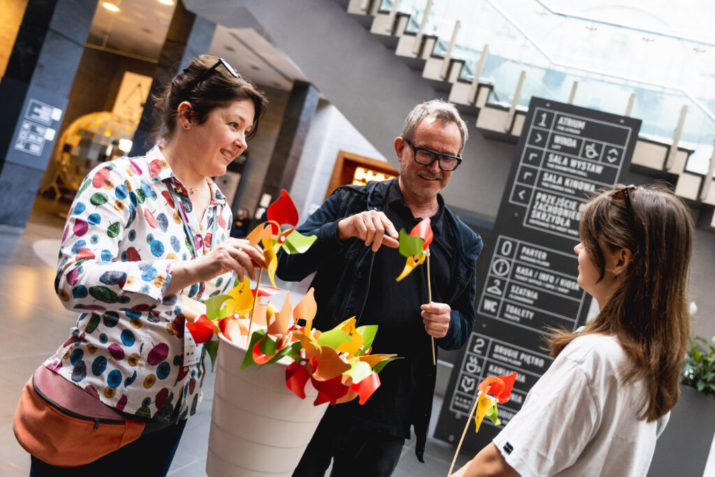 Trzy osoby stoją w holu poznańskiego Zamku. Obok nich w pojemniku kolorowe wiatraki.