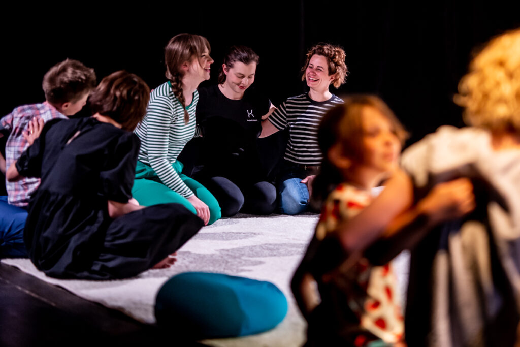 Na scenie rozłożona jest dywan. Na nim siedzi grupa dzieci i dorosłych.