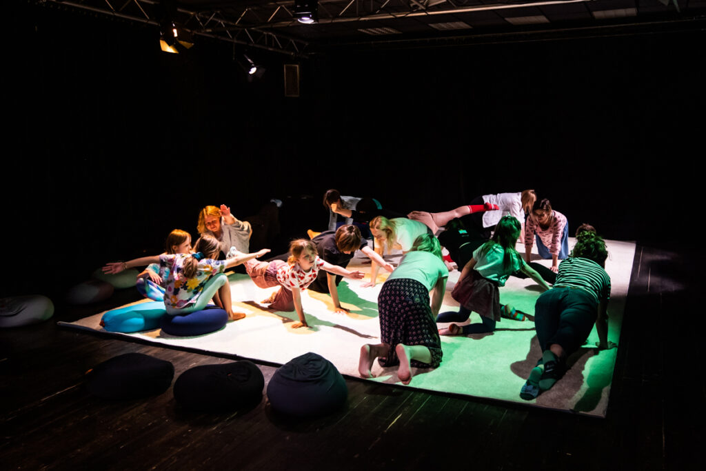 Na scenie na rozłożonym dywanie grupa dzieci i dorosłych klęczy unosząc ręce lub nogi. Pada na nich światło.