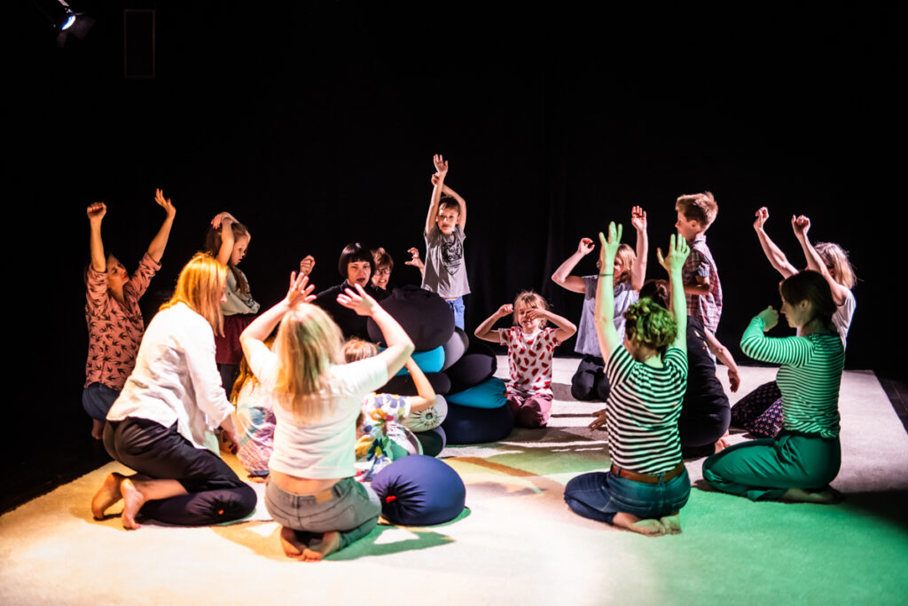Grupa dzieci i dorosłych siedzi na dywanie rozłożonym na scenie. Unoszą ręce do góry.