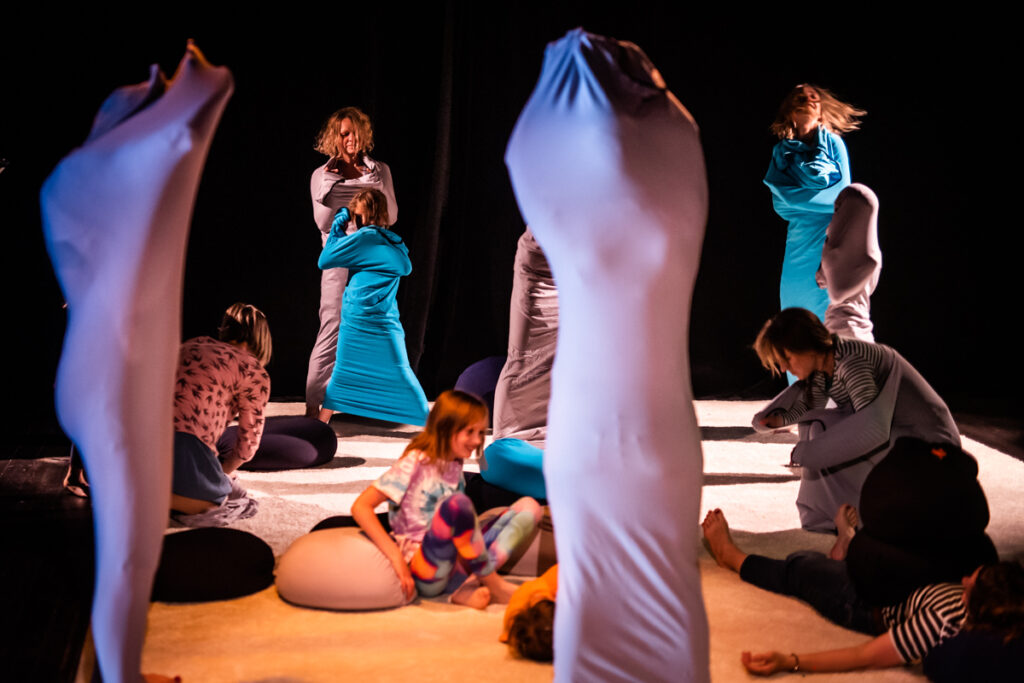 Na scenie rozłożony dywan. Na nim grupa dzieci i dorosłych ubiera się w ciasne kostiumy.