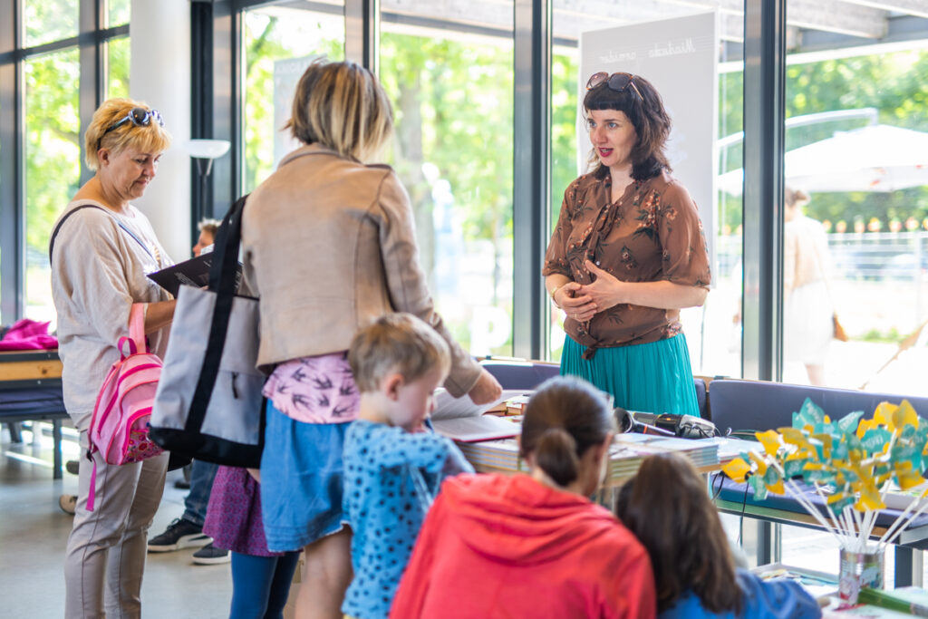 Grupa dzieci i dorosłych stoi przed ladą w budynku Sceny Wspólnej. Za ladą stoi kobieta.