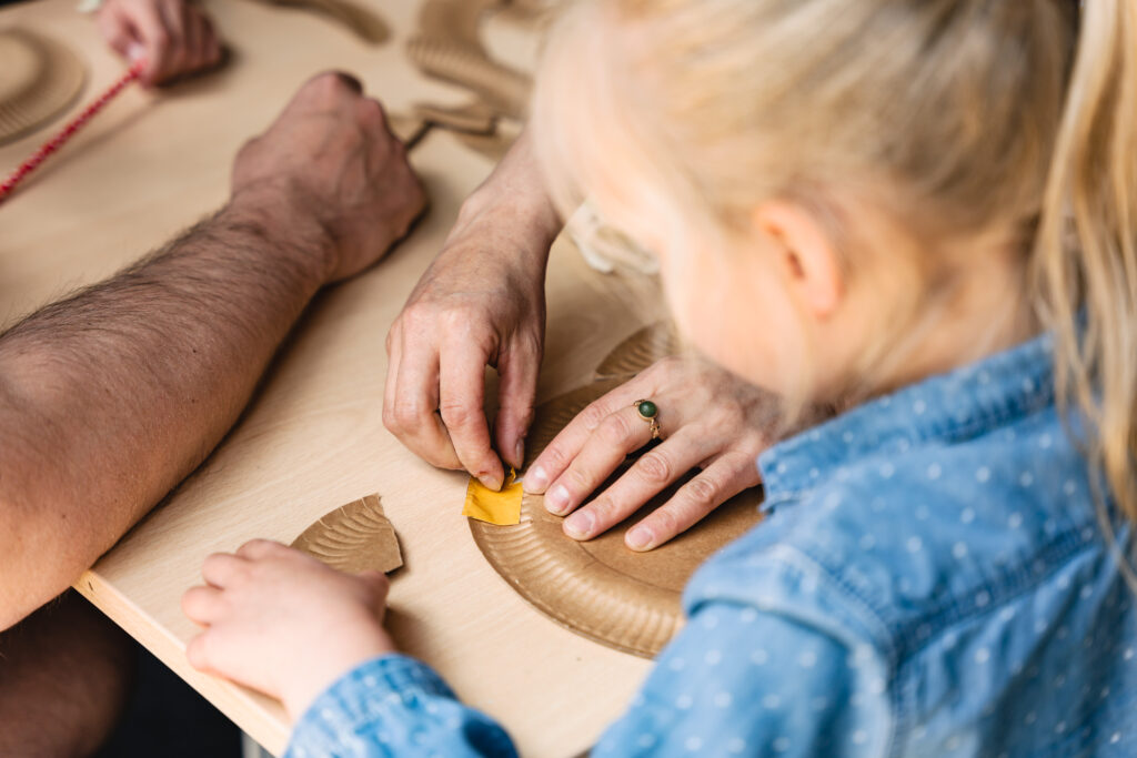 Dziewczynka kleni papierowy talerzyk. Pomaga jej ktoś dorosły.