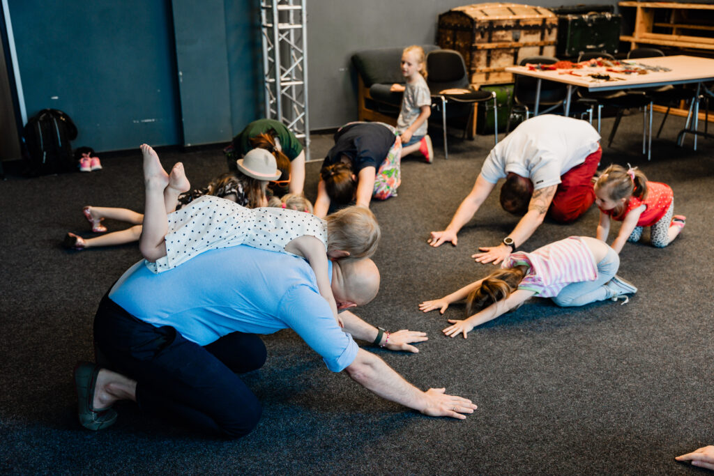 Warsztaty. Grupa dzieci i dorosłych klęczy na podłodze. Na przedzie dziewczynka leży na plecach mężczyzny.