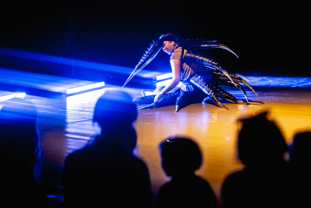 Na przedzie widownia. W tle aktorka siedzi na scenie. Ma założone mnóstwo długich ogonów wyrastających z różnych części ciała.
