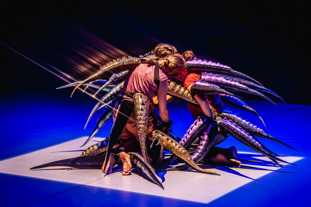 Trójka aktorów na scenie. Klęczą zwróceni do siebie. Z ich ciała wyrasta mnóstwo długich ogonów.
