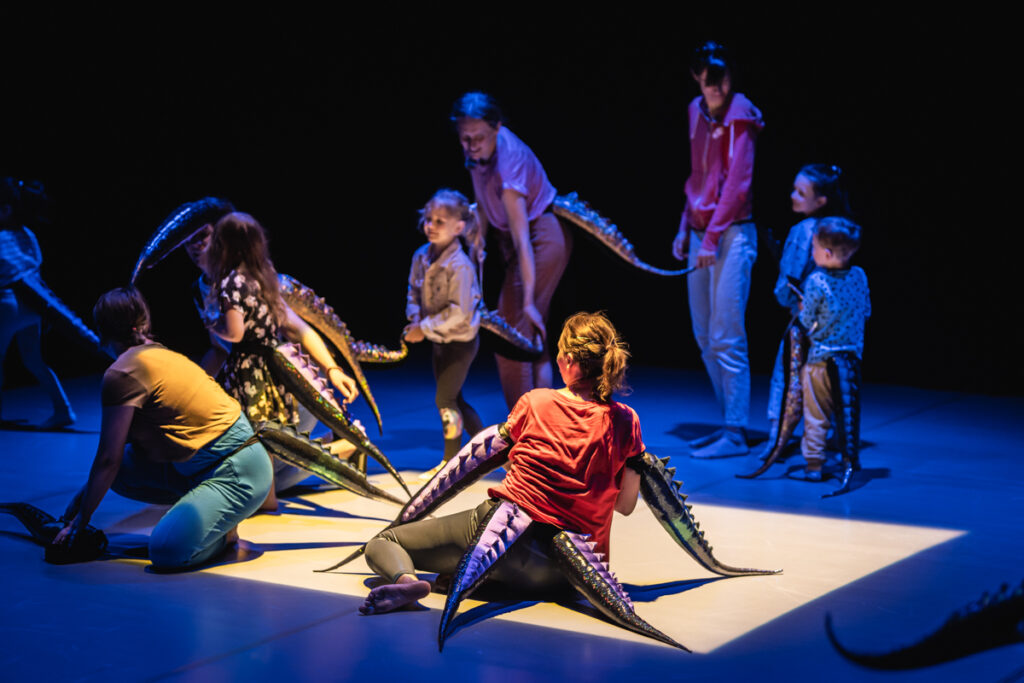 Na scenie grupa dzieci i dorosłych. Wszyscy mają przywiązane długie ogony.