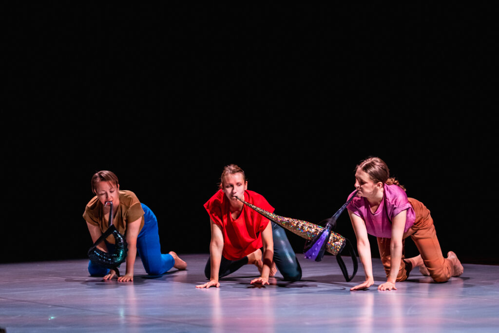 Na scenie. Trzy aktorki siedzą na czworaka. W ustach trzymają długie kolorowe obiekty.