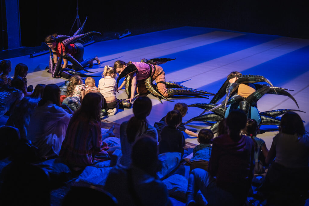 Na scenie trzy aktorki poruszają się na czworaka. Mają do siebie przywiązanych mnóstwo długim ogonów. Na przedzie widownia. Aktorkom przyglądają się z zaciekawieniem dzieci siedzące na scenie.