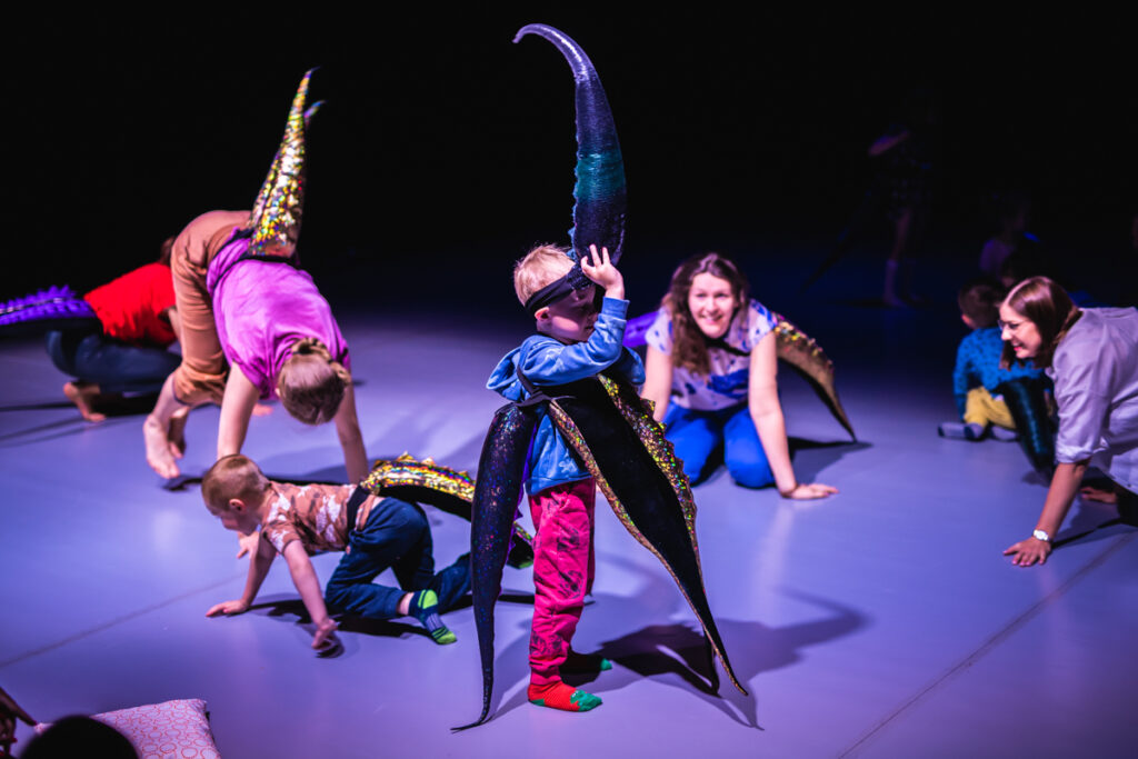 Grupa dzieci i dorosłych na scenie. Mają przywiązane długie ogony. Na środku stoi chłopiec, który ma ich kilka. / A group of children and adults on stage. They have long tails tied to them. In the middle stands a boy who has several of them.