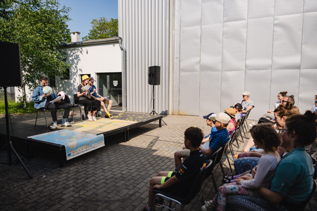 Po lewej scena, na której siedzą aktorz. Jeden z nich trzyma w rękach globus. Na scenie z przodu leży mapa. Po prawej stronie widownia.