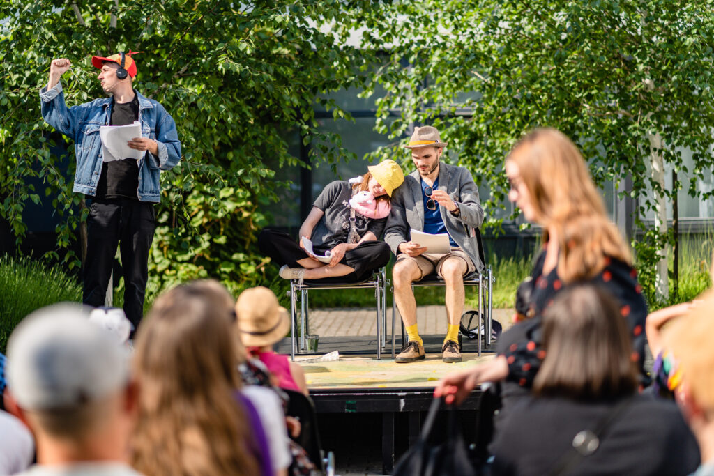 Z przodu grupa osób. W tle aktorzy na scenie. Dwójka siedzi na krzesłach. W rękach mają skrypt. Po lewej aktor stoi ze skryptem w rękach.