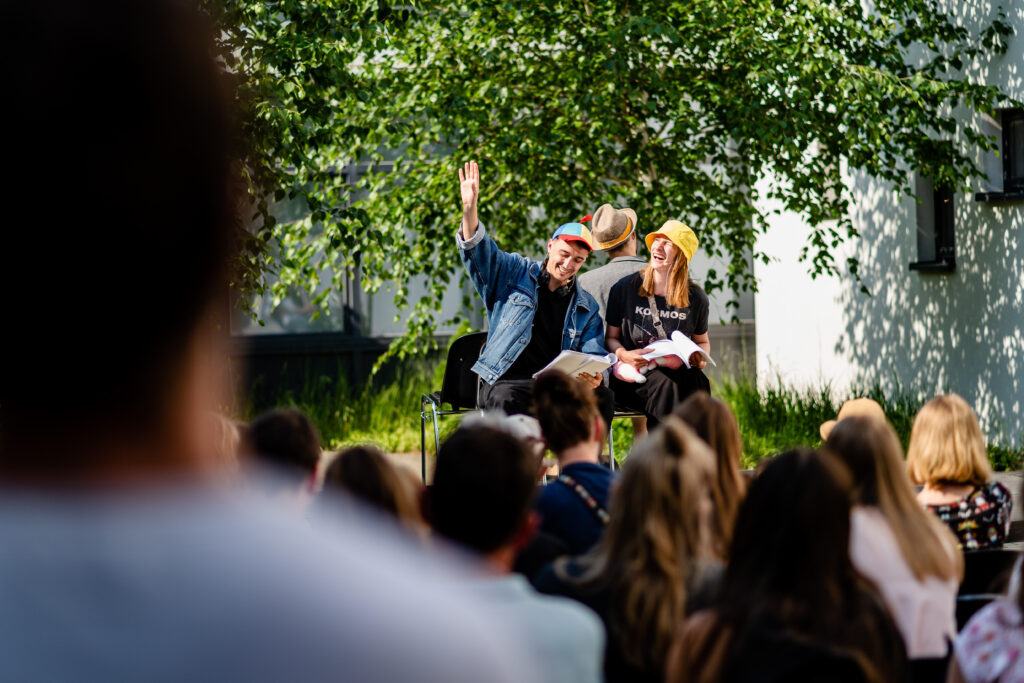 W tle na scenie grupa trójka aktorów siedzi na krzesłach. W rękach mają skrypty. Z przodu siedzi widownia.