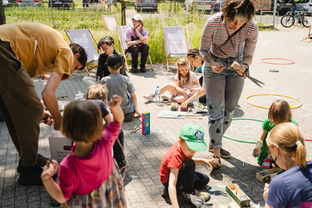 Grupa dzieci i dorosłych. Mają przybory do malowania. Na ziemi leżą kolorowe skrzyneczki.