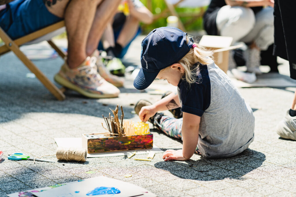 Dziecko siedzi na ziemi. Obok niego leży kolorowa skrzynka, do której wtyka patyczki.
