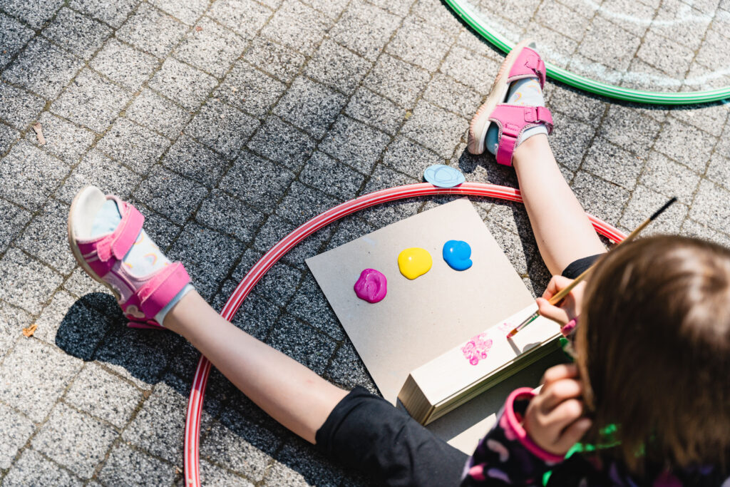 Na ziemi siedzi dziecko. Przed sobą ma karton z farbami. Maluje na drewnianej skrzyneczce.