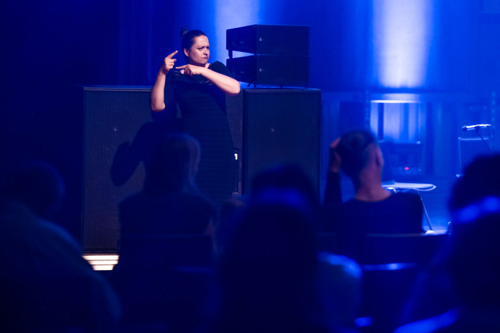 W tle widzoczna kobieta używające języka mogowego skierowana w stronę widowni.